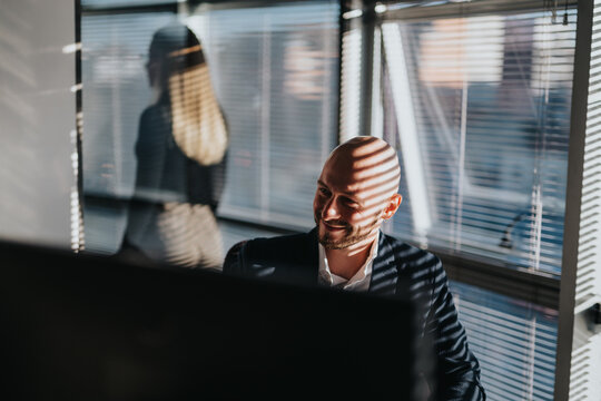 A businessperson sits at a desk in an office, smiling while working at a computer; sunlight creates striped shadows across the scene.