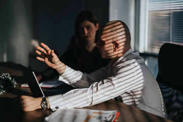 Fototapeta premium A business person gestures while discussing at a laptop. Another business employee listens in a sunlit, striped-blind office.