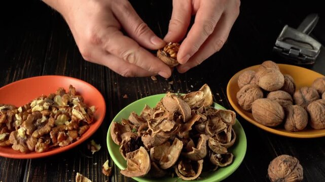 Hands cracking open walnuts, separating shells from nuts, with bowls of nuts and shells arranged on a dark wooden table