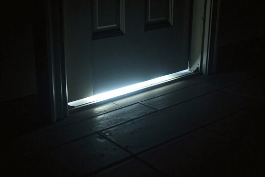 Faint glow under a room door, only light in the pitch darkness late at night. Concept of nefarious activity behind close doors, working late, afraid of the dark, turning lights on going to sleep.