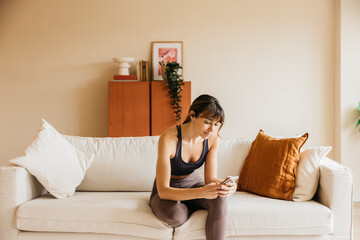 Woman sitting on a couch in comfortable yoga wear, browsing her smartphone