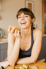 Woman in sportswear enjoying a fresh orange in her kitchen, showing healthy eating and happiness
