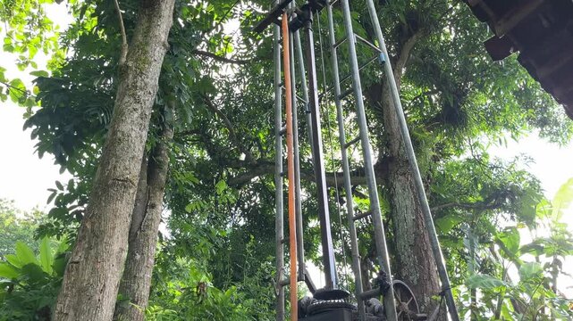 Water well drilling rig positioned beside a large tree in a tropical rural area, showing interaction between heavy equipment and natural surroundings. The footage represents sustainable development an