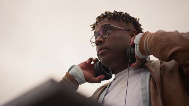 black man near console focused low angle cinematic shot headphones resting equipment silhouette foreground leather jacket glasses serious producer stance ambient urban atmosphere dramatic sky