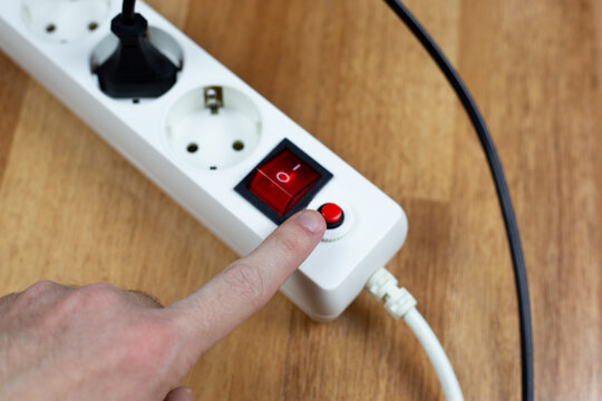 Hand approaches circuit breaker reset button and red master switch of white power strip on wooden desk. This emphasizes user interaction, safety checks, and manual control over power