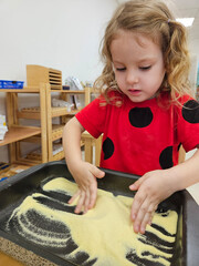 Little girl is playing with sand in a pan. She is wearing a red shirt with black polka dots