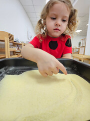 Little girl is playing with sand in a tray. She is wearing a red shirt and black polka dots