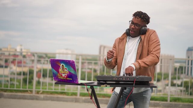 black dj rooftop upbeat mix energetic performance with active mixing, turning knobs and cueing tracks on controller and turntable, wearing brown jacket and headphones, colorful laptop sticker, urban
