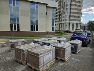 Pile of boxes with pavement bricks are stacked on a cement floor. The boxes are wrapped in plastic and are stacked in a row near entrance in the courtyard of the house in residential complex