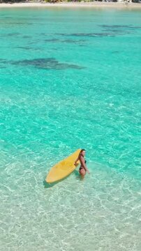 Woman leisurely strolls with a yellow paddle board in clear blue waters on a sunny day, creating a tropical paradise vibe perfect for a dreamy vacation