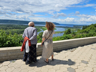 Two women are standing on a stone walkway overlooking a lake or river Volga. One of the senior women is wearing a red coat