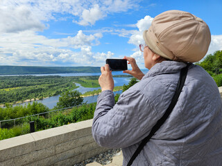 Woman senior is taking a picture of a beautiful landscape. She is wearing a hat and a gray jacket