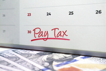 Calendar with the phrase Pay Tax handwritten in red ink over US hundred dollar bills. Concept for tax deadlines, fiscal responsibility, and government revenue obligations.