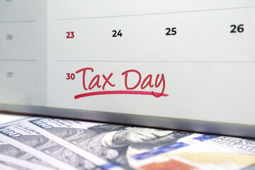 Calendar with the phrase Tax Day handwritten in red ink over US hundred dollar bills. Concept for the annual tax filing deadline, financial preparation, and IRS submission dates.