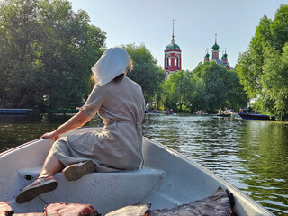 Woman in a white hat is sitting in a boat on a lake. The boat is near a church