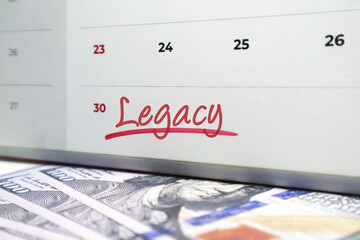 White spiral calendar with the word Legacy handwritten in red ink, lying on top of scattered US hundred dollar bills. Concept for inheritance.
