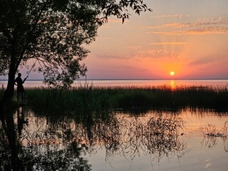 Man is fishing in a lake. The sun is setting in the background. The water is calm and the sky is orange