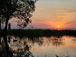 Man is fishing in a lake. The sun is setting in the background. The water is calm and clear
