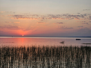 Boat is in the water near the shore. The sky is orange and pink. The water is calm