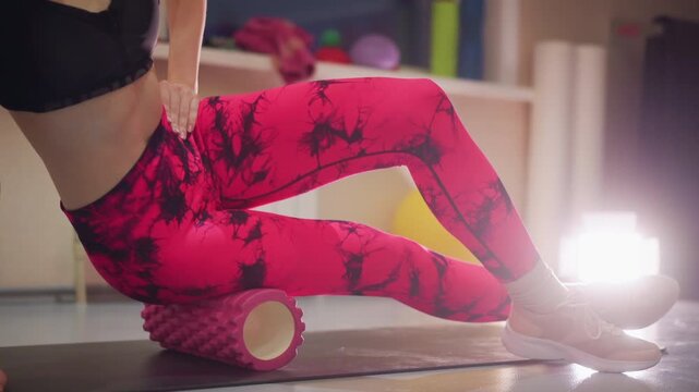 Closeup legs foam roller hip release, black sports bra pink patterned leggings, indoor studio mat textured roller under IT band slow controlled movement muscle massage warm backlight calm focused
