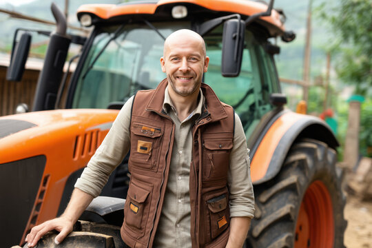 Portrait of Caucasian middle aged man smiling while standing in front of large tractor outdoors, showing confidence and approachability in agricultural or farming environment