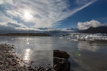 Bright sunlight glows across Jökulsárlón glacier lake as blue and white icebergs drift beneath Southern Iceland’s towering mountains and soft clouds mirrored in the still midday water © lightcaptured