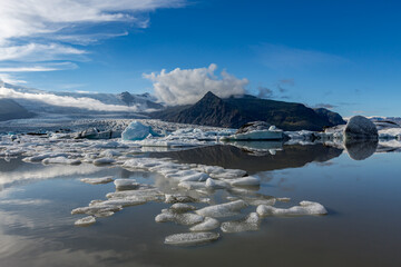 Bright sunlight glows across Jökulsárlón glacier lake as blue and white icebergs drift beneath Southern Iceland’s towering mountains and soft clouds mirrored in the still midday water © lightcaptured