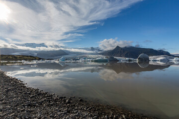Bright sunlight glows across Jökulsárlón glacier lake as blue and white icebergs drift beneath Southern Iceland’s towering mountains and soft clouds mirrored in the still midday water © lightcaptured