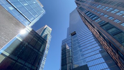 Modern skyscrapers with glass and brick facades are reaching up into a deep blue sky, featuring sunlight reflecting off the building surfaces, creating a vibrant urban scene