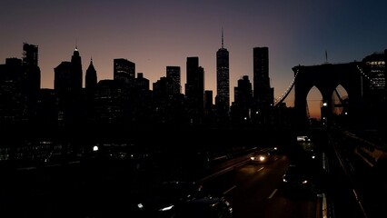 New york city financial district skyline with iconic one world trade center and brooklyn bridge arches silhouetted against the colorful evening sky, cars circulating on the road below © DawDunia