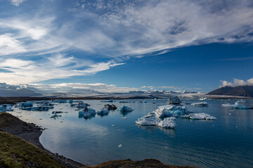 Blue and white icebergs drift across Jökulsárlón glacier lake as warm afternoon light settles over Southern Iceland’s sweeping glacier and rugged mountains rising quietly beyond the still lagoon © lightcaptured