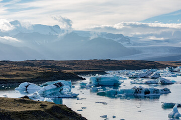 Blue and white icebergs drift across Jökulsárlón glacier lake as soft evening light settles over Southern Iceland’s rugged hills and the distant glacier rising quietly beyond the mirrored water © lightcaptured