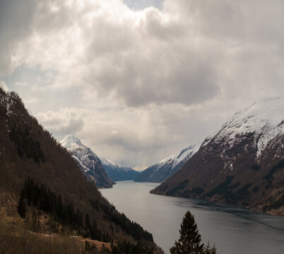  Fjordtal mit t&uuml;rkisem See, schneebedeckten Gipfeln, bewaldeten H&auml;ngen und bew&ouml;lktem Himmel &ndash; typisch f&uuml;r Fr&uuml;hling in Westnorwegen