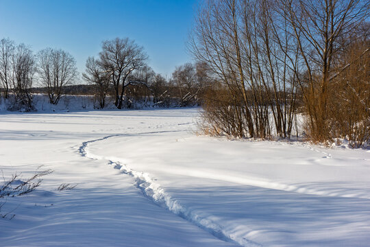 Footprints wind across a pristine snowscape towards a frozen riverbank under a bright blue sky