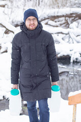 A young Caucasian man with glasses carries two water containers in a snowy landscape. He wears a black coat and a blue beanie, standing near a stream.