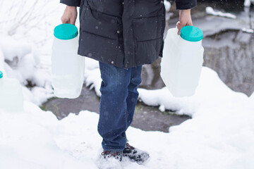 A young Caucasian man with glasses carries two water containers in a snowy landscape. He wears a black coat and a blue beanie, standing near a stream.