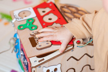 A small child plays with a colorful wooden toy house. The child has blond hair and is wearing a beige suit. The busy board toy has gears and various shapes.