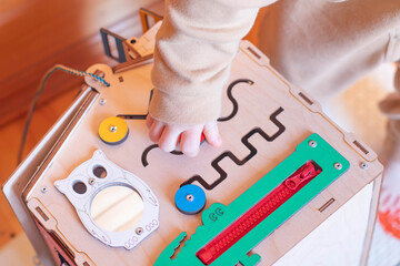A small child plays with a colorful wooden toy house. The child has blond hair and is wearing a beige suit. The busy board toy has gears and various shapes.