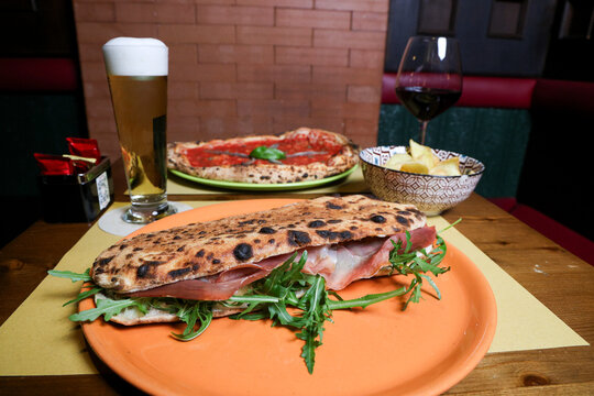 Close-up view of a casual dining table featuring a gourmet burger, a sandwich, a Panuozzo, a glass of draft beer and red wine in a modern restaurant or pub setting.