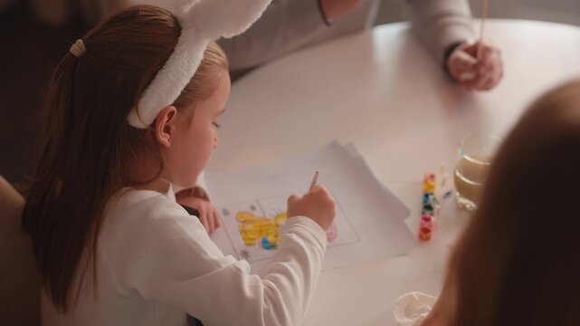 Adorable Young Girl in Bunny Ears Coloring an Easter Picture with Paints at Home