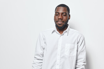 Obraz premium African American man with dark skin wearing white shirt standing with eyes closed against white background. Portrait of calm black male with beard and short hair in casual style.