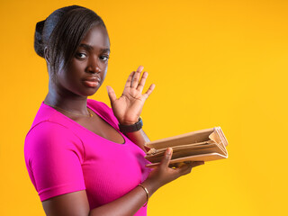 A woman in a vibrant pink top poses against a bright yellow background, holding open an empty tan wallet with a skeptical expression. Her hand is raised in a gesture of disbelief or shrug