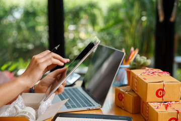 Close up of business woman hands using digital tablet and stylus pen to check online orders. SME entrepreneur working with parcel boxes on wooden table with green garden background.