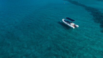 High-angle shot of a white rigid inflatable boat (RIB) with a black bimini top, anchored in crystal-clear turquoise sea water. The rocky seabed is clearly visible through the shallow, calm water. © Stefano Tammaro