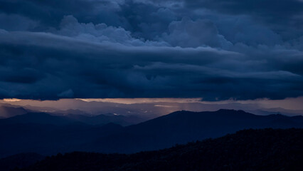 Dramatic storm clouds with heavy rain over mountain silhouette during sunset. Dark sky contrast with golden sunlight background.