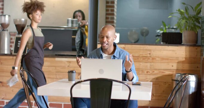 African American man waving on call, barista crossing behind chair, typing on silver laptop at cafe