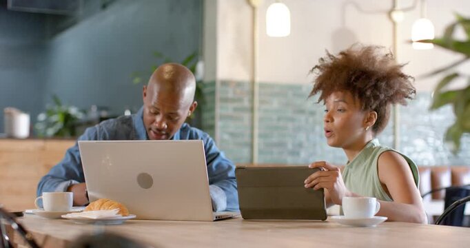 African American colleagues at cafe woman showing tablet speaking while man typing laptop working