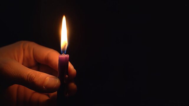 A human hand gently holds a lit purple candle, its bright flame illuminating the fingers and the wax against a completely dark background.