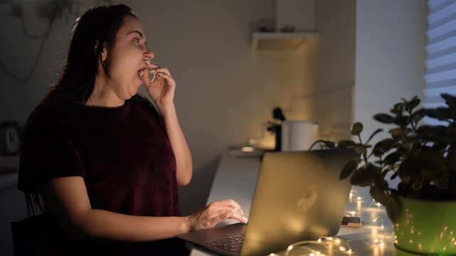 Tired woman in pajamas yawning in front of laptop computer, working late at night in the kitchen.