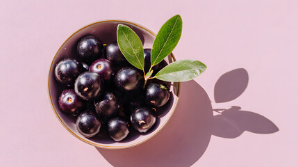 Bowl of glossy dark purple berries with green leaf on pastel pink background casting soft shadow, minimal food still life composition